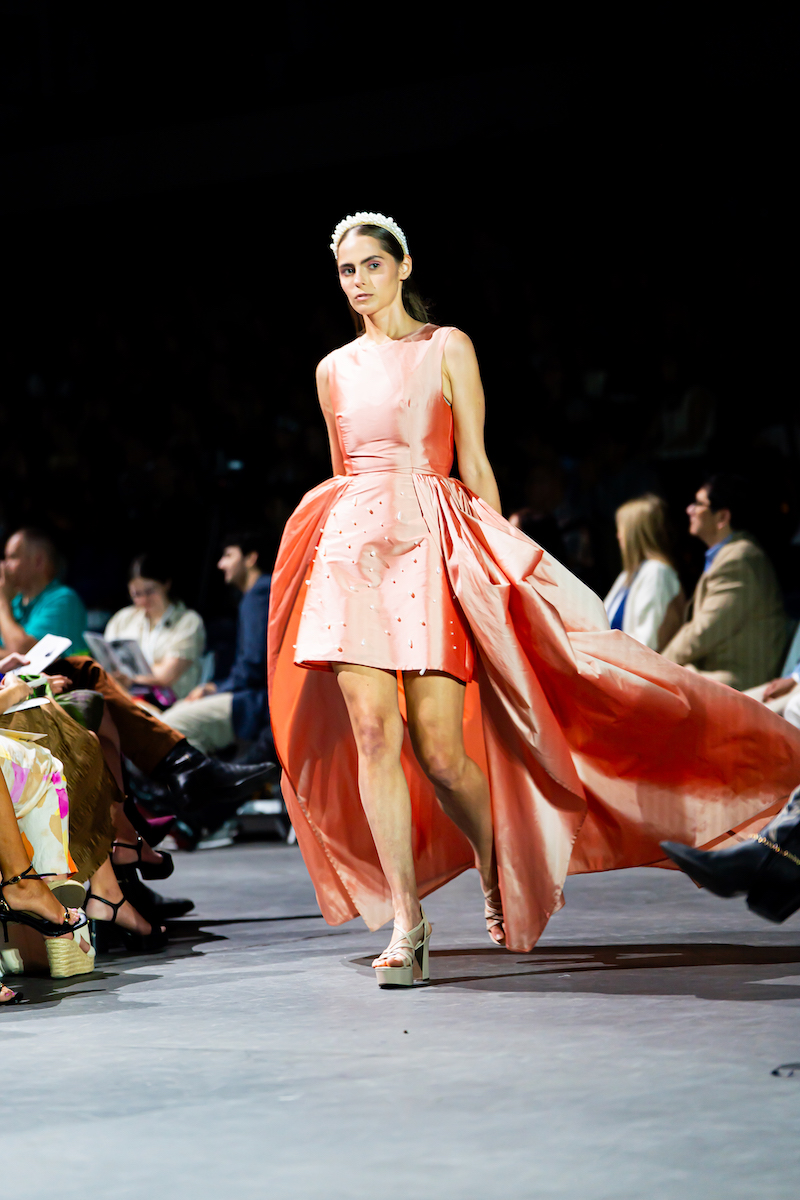 Model in coral high-low dress with white headband walking a fashion runway.