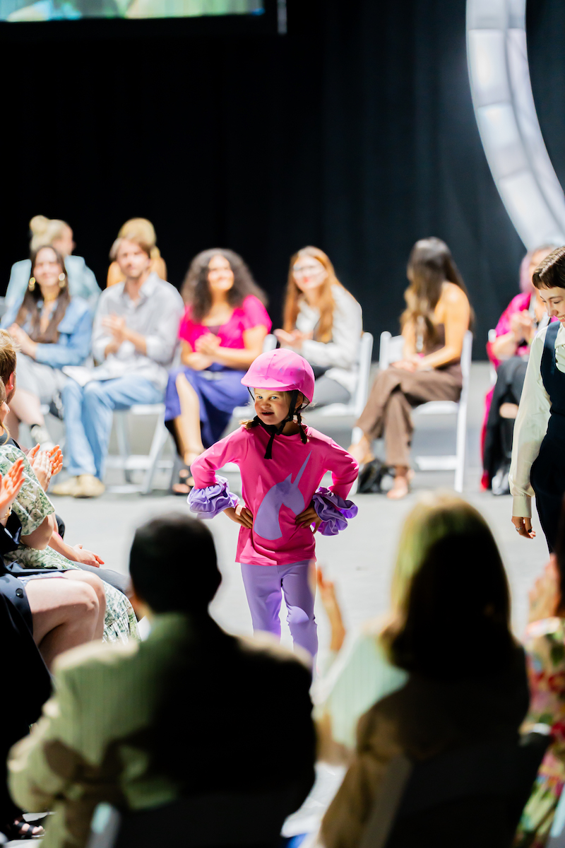Young model in pink and purple adaptive outfit walks runway confidently as audience claps and smiles.