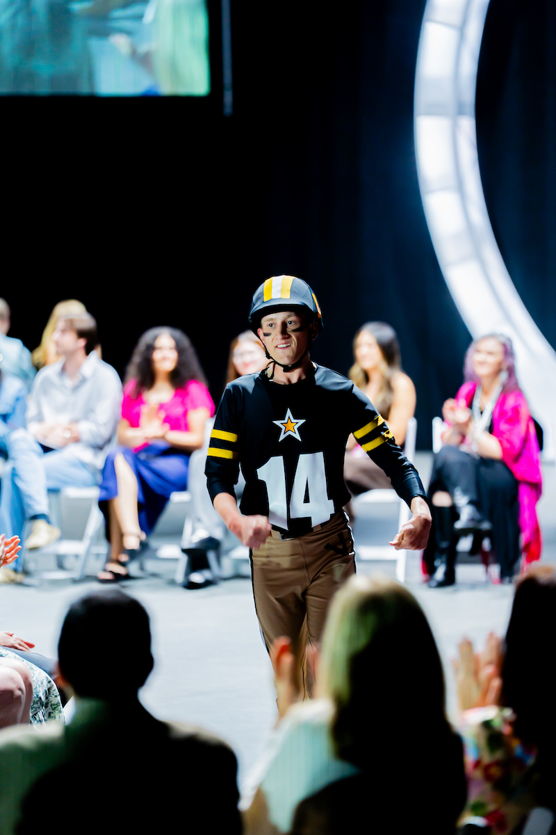 Young model in star-themed jersey and helmet walks confidently down the runway to audience applause.