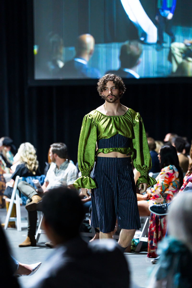 Male model wears vibrant green puffed-sleeve top and striped shorts during O'More fashion show.