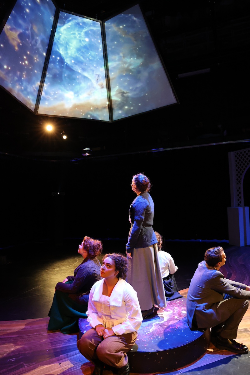 actors sitting on a stage looking up at a multicolored nebula projected on an LED screen