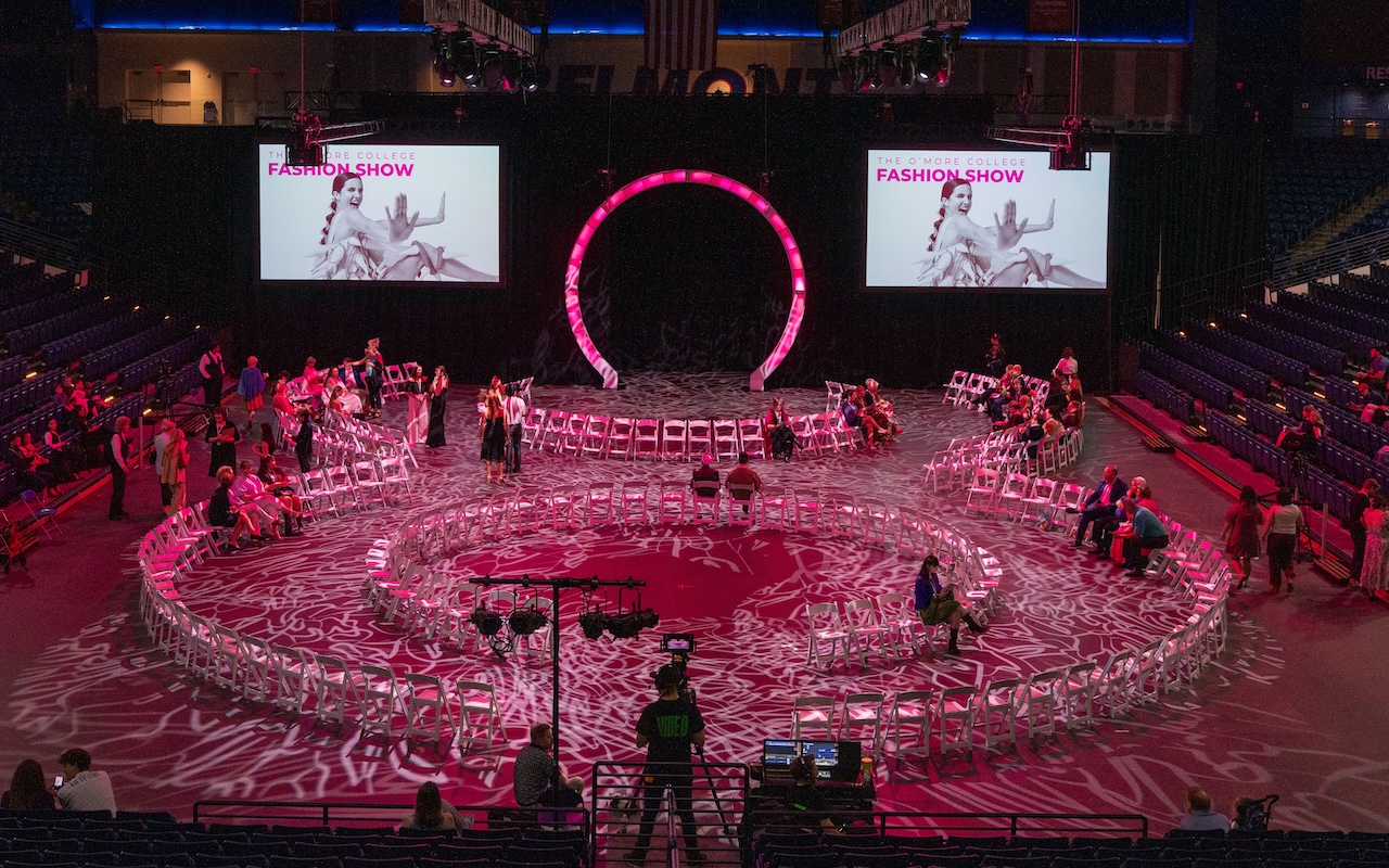The 2025 O’More Fashion Show at Belmont University features a dramatic circular runway, glowing pink lighting, and large display screens with event graphics as guests begin to arrive.