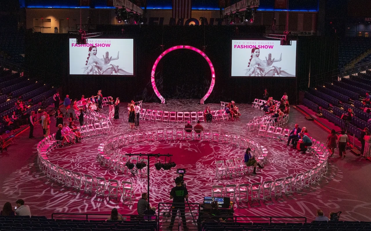 The 2025 O’More Fashion Show at Belmont University features a dramatic circular runway, glowing pink lighting, and large display screens with event graphics as guests begin to arrive.