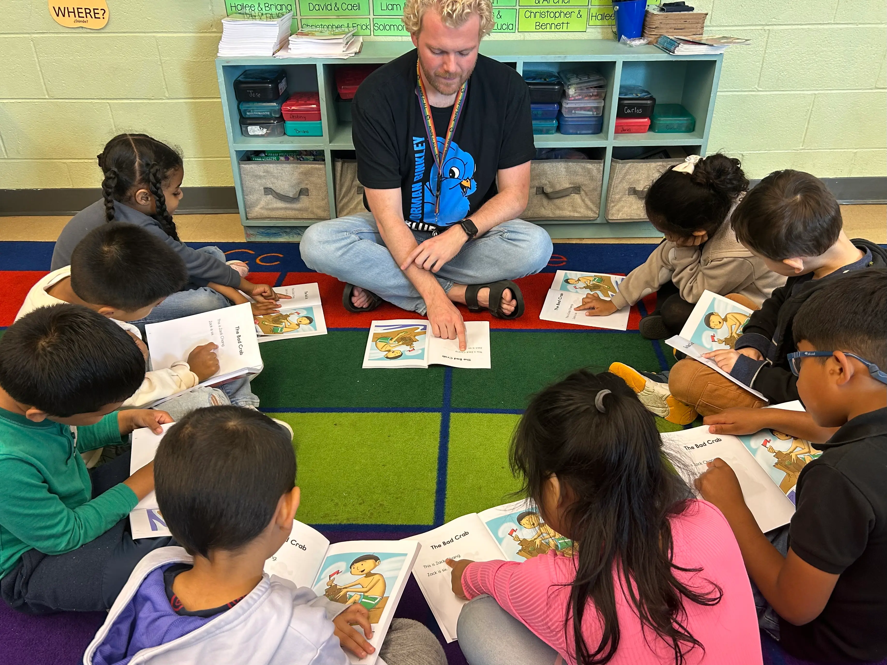 A teacher with blond hair wearing a black t-shirt with a blue design sits cross-legged on a colorful classroom rug, leading a reading group with elementary students who are holding open picture books titled "The Red Crab."