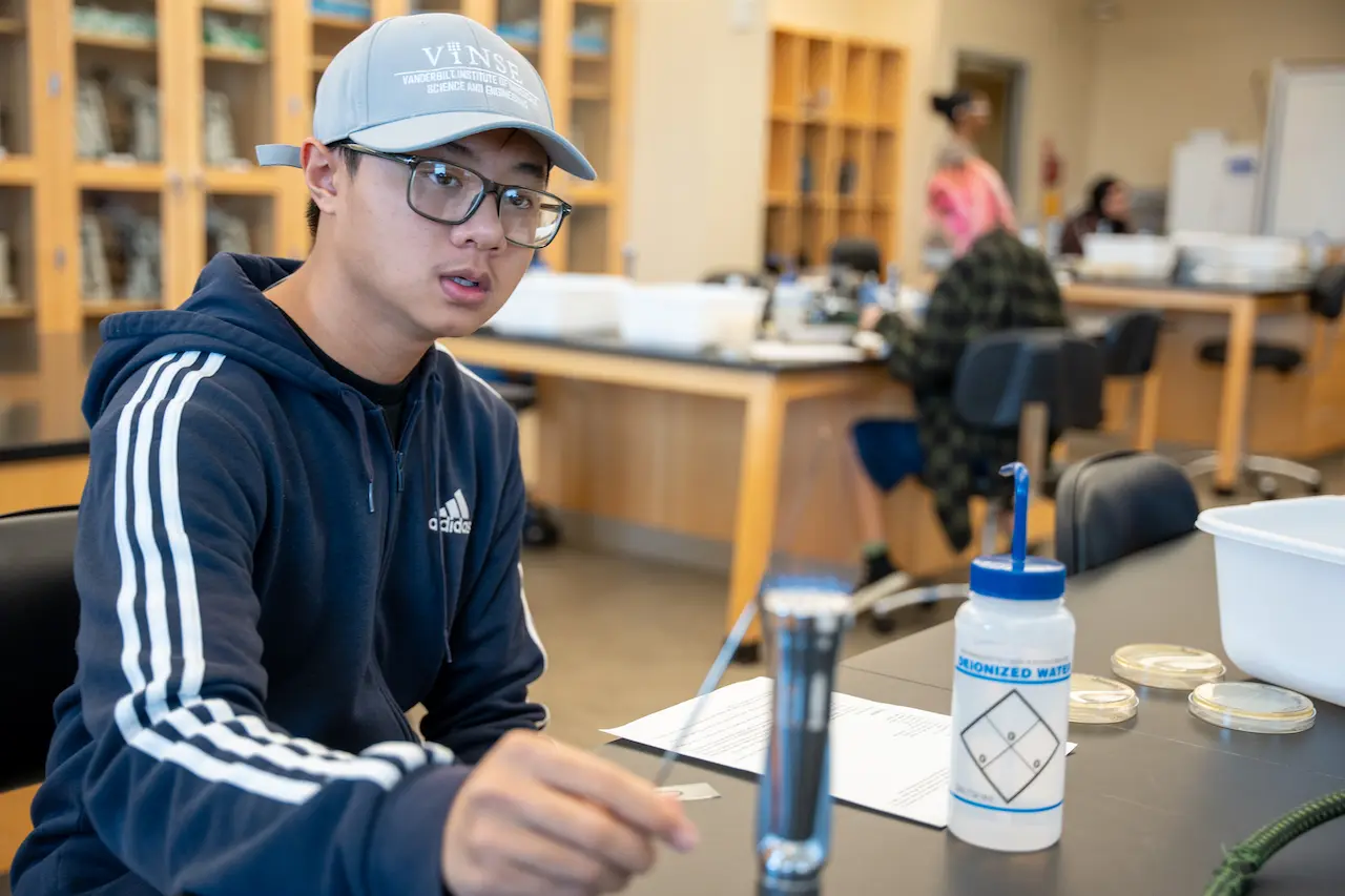 A student wearing a blue Adidas hoodie and a VINSE cap sits at a lab table with scientific equipment, including petri dishes and a deionized water bottle.