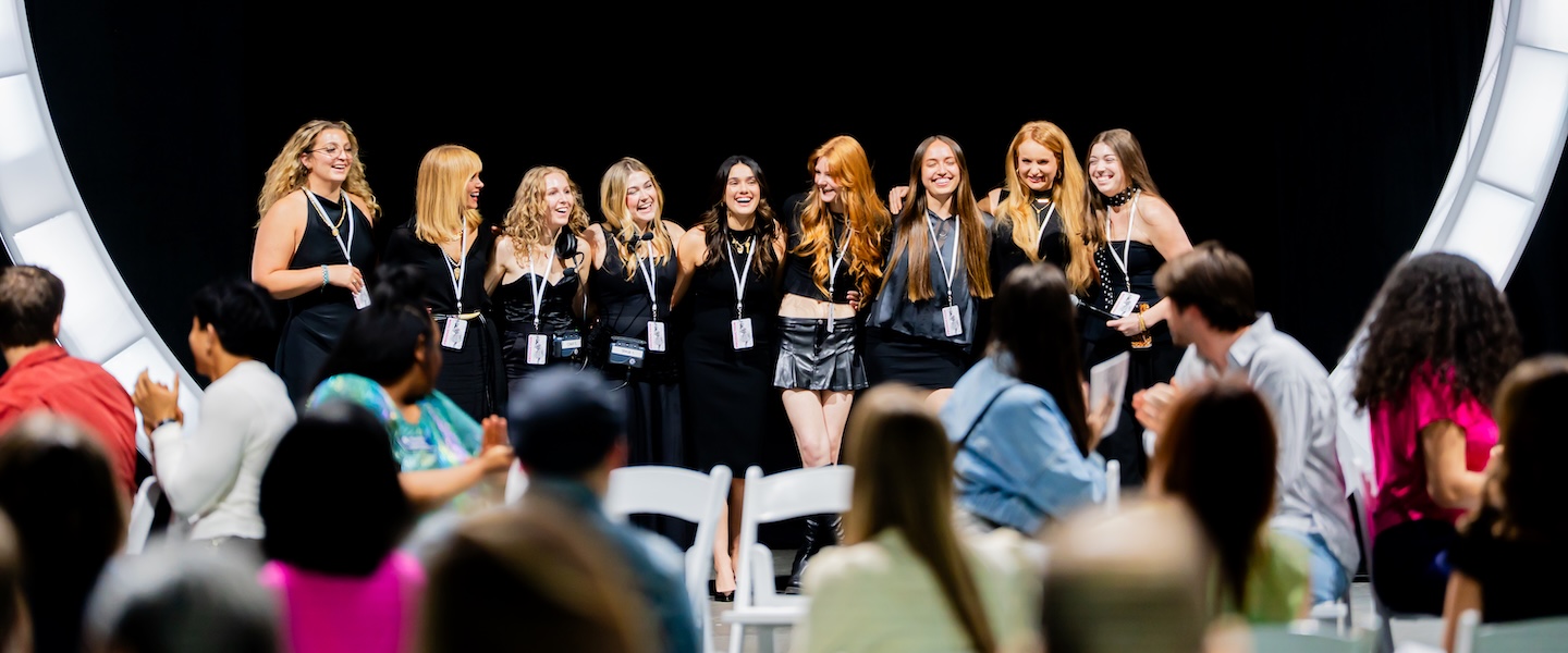 Fashion merchandising students take the stage at the 2025 O'More Fashion Show, smiling in coordinated black outfits as the audience applauds.