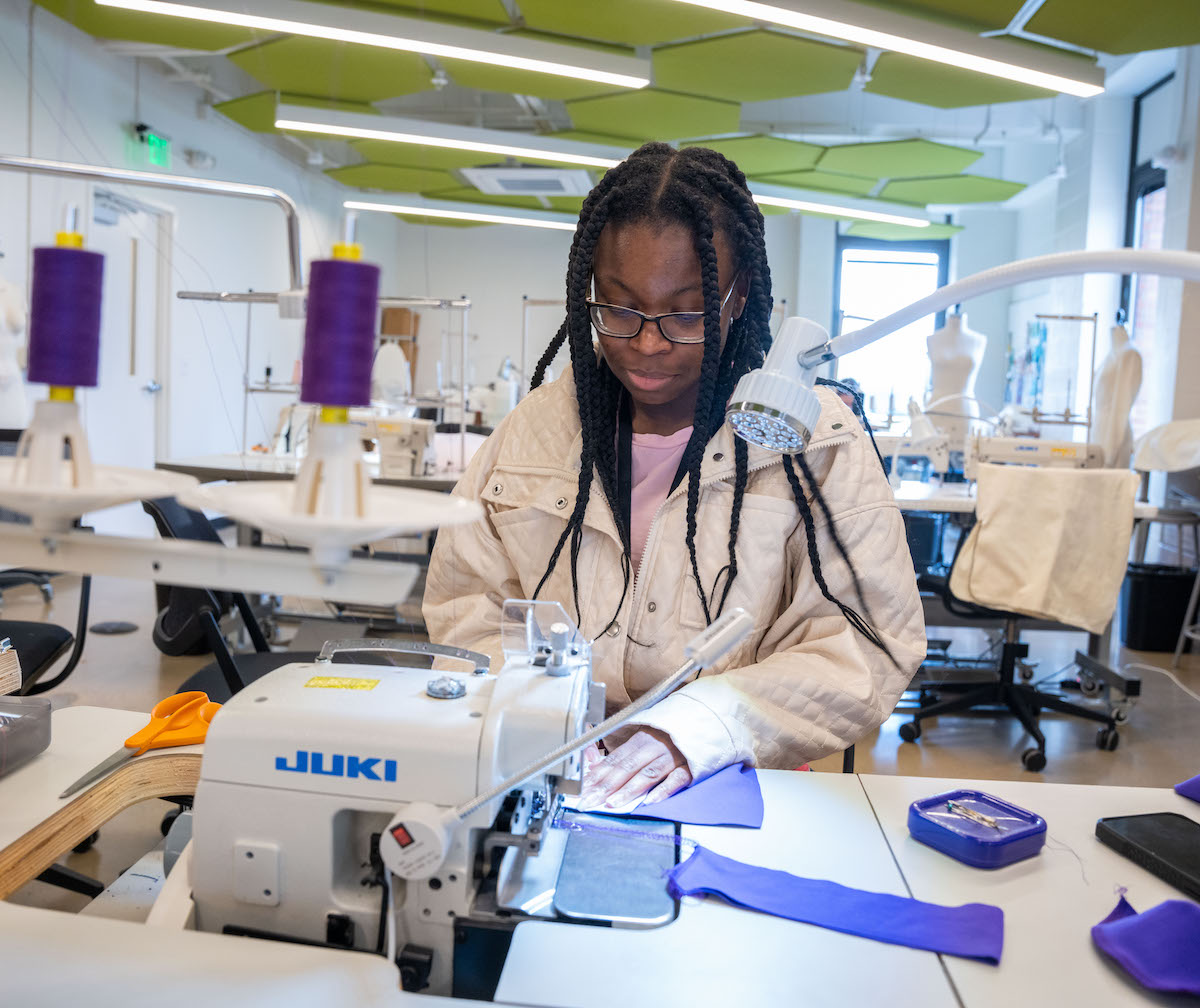 Student Trisha works with a sewing machine in a fashion design lab at Belmont University, stitching a piece of purple fabric surrounded by tools and thread spools.