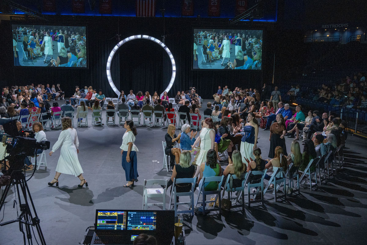 Models walk the figure-eight runway during the 2025 O’More College Fashion Show as audience members look on and applaud; large screens display a live feed of the show, and a backstage control station is visible in the foreground.
