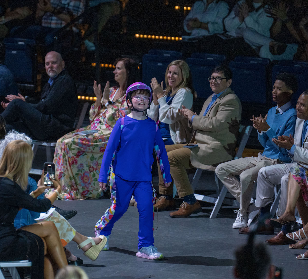 Ephraim walks confidently down the runway at the 2025 O’More College Fashion Show wearing a bright blue adaptive outfit and matching helmet, while the audience applauds.