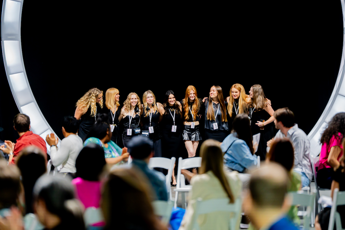 Fashion Event Team dressed in black poses together on stage under a glowing circular arch during the 2025 O’More College Fashion Show, as the audience applauds.