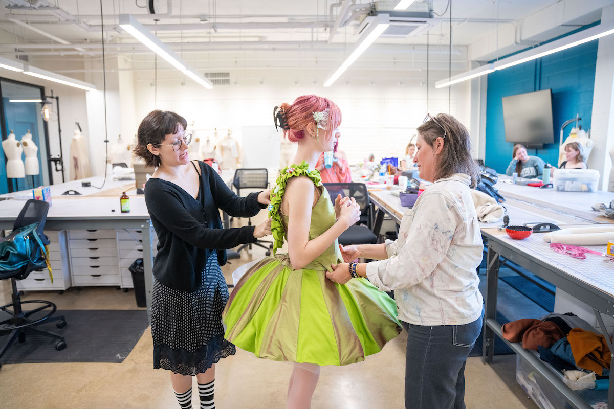 A faculty member and student assist a model during a dress fitting in Belmont’s fashion design studio, adjusting a bright green structured garment while others work in the background.