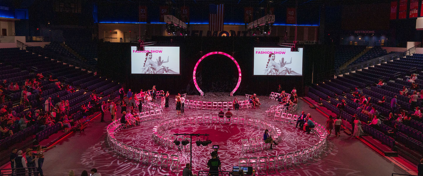 Overhead view of the 2025 O’More College Fashion Show, featuring a glowing figure-eight runway with white chairs arranged around it in a large indoor arena, illuminated with pink lighting and decorative projections.