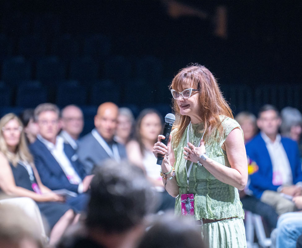 Jamie Atlas speaks into a microphone while addressing the audience at the 2025 O’More College Fashion Show, standing in front of a seated crowd and wearing a light green outfit with a fashion show badge.