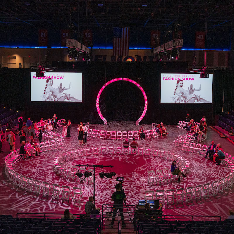Overhead view of the 2025 O’More College Fashion Show, featuring a glowing figure-eight runway with white chairs arranged around it in a large indoor arena, illuminated with pink lighting and decorative projections.