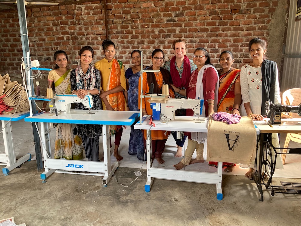 A group of women from Harvest Frontiers stand behind sewing machines in a workshop with brick walls. 