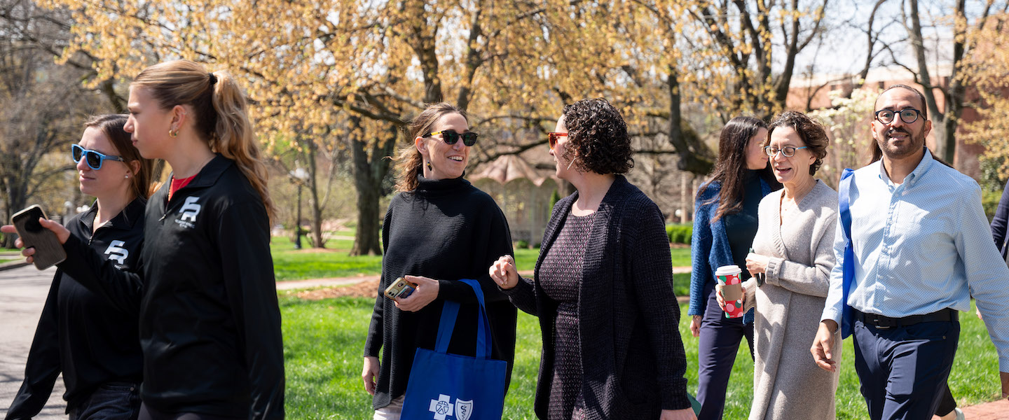Belmont community walking together outside on campus, smiling and talking on a sunny day with trees in the background.