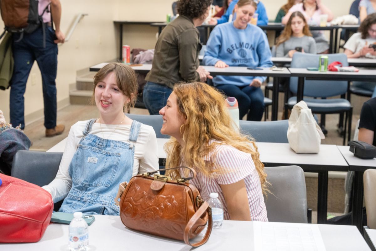 Two girls sit at a desk in a classroom and smile at the 2025 Belmont Literary Journal launch party