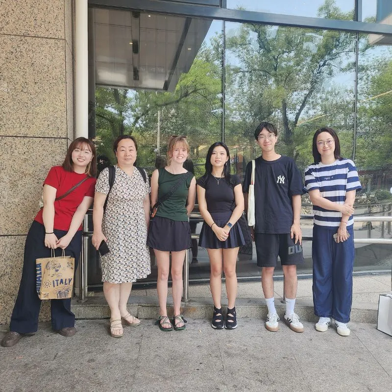 Group of six people standing together outside a building with large glass windows, smiling and posing for the camera.