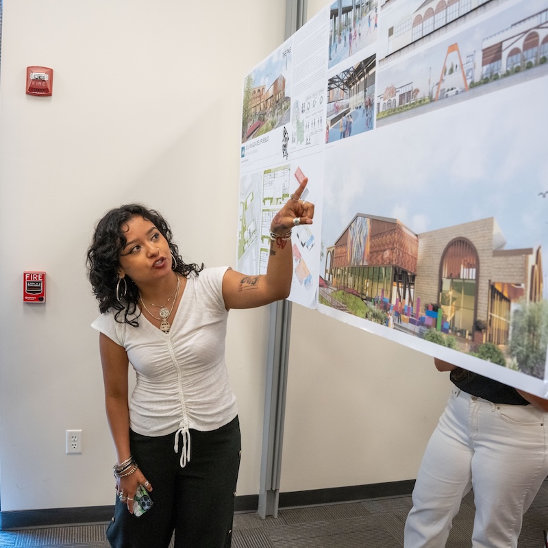 Student gestures while presenting architectural design board featuring colorful renderings and site plans.