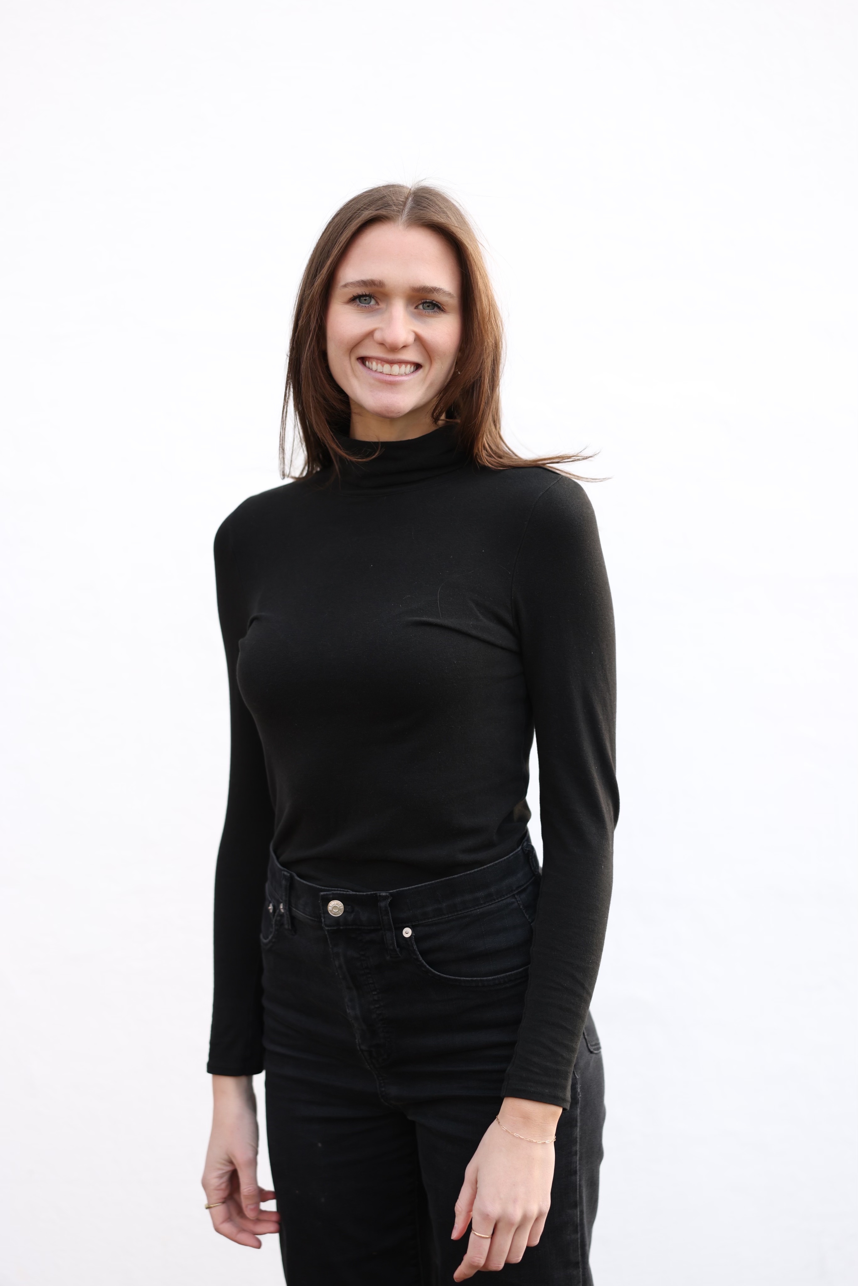 Rachel Moore standing and smiling at the camera, wearing a black turtleneck and black jeans, in front of a plain white surface.
