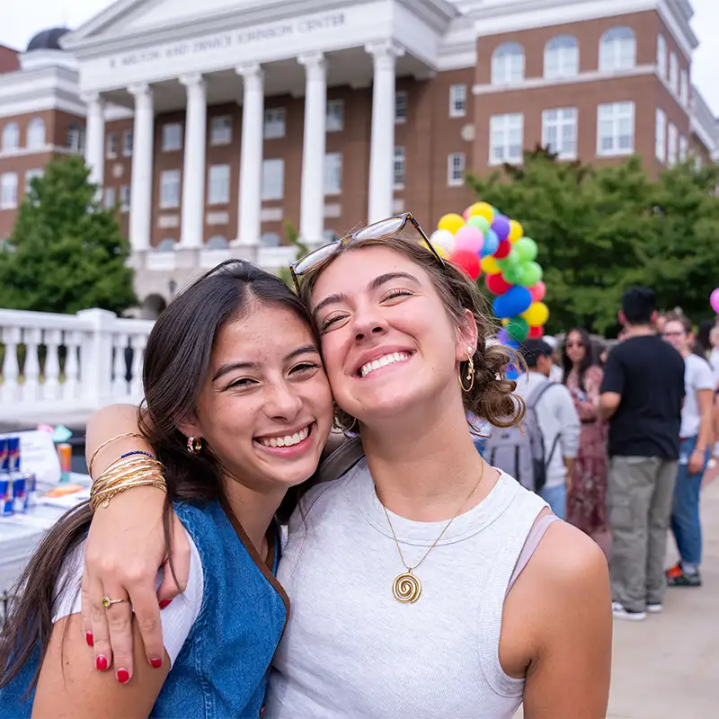 Two young women are smiling and embracing each other in front of a large building with columns. The woman on the left has long, dark hair and is wearing a denim vest, while the woman on the right has light brown hair styled in loose curls and is wearing a light gray shirt. In the background, there are colorful balloons and a crowd of people enjoying an outdoor event.