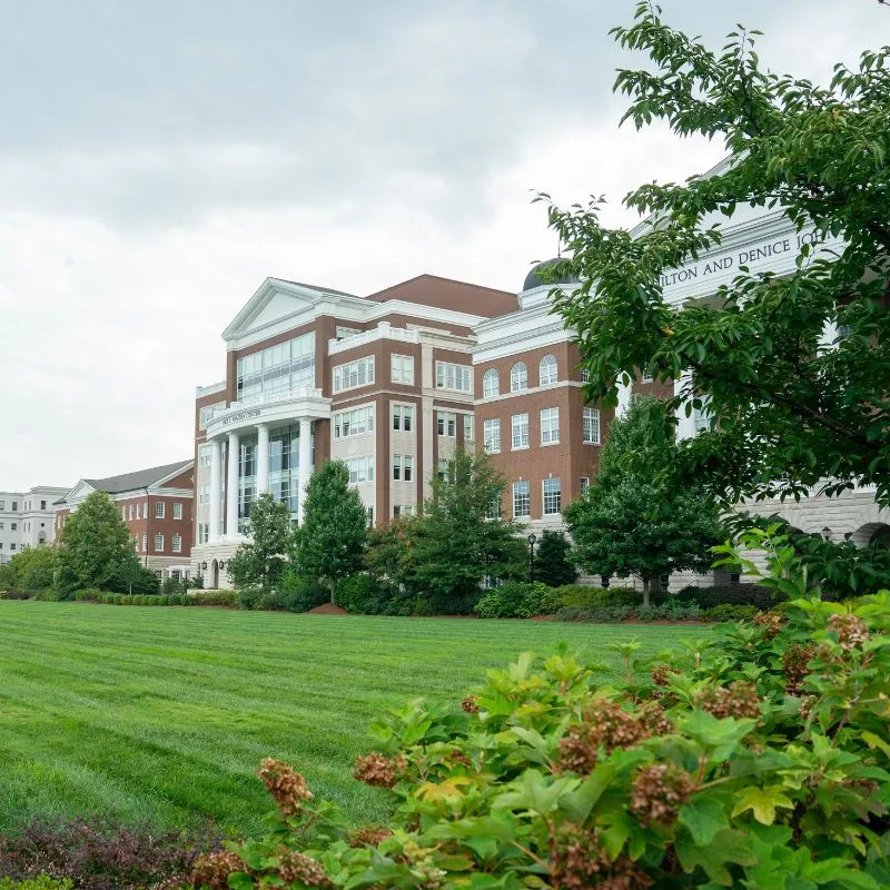 A view of the Jack C. Massey building on the Belmont University campus, featuring a large glass entrance and brick facade. The surrounding landscape includes well-maintained lawns and trees, under a cloudy sky, creating a serene educational environment.