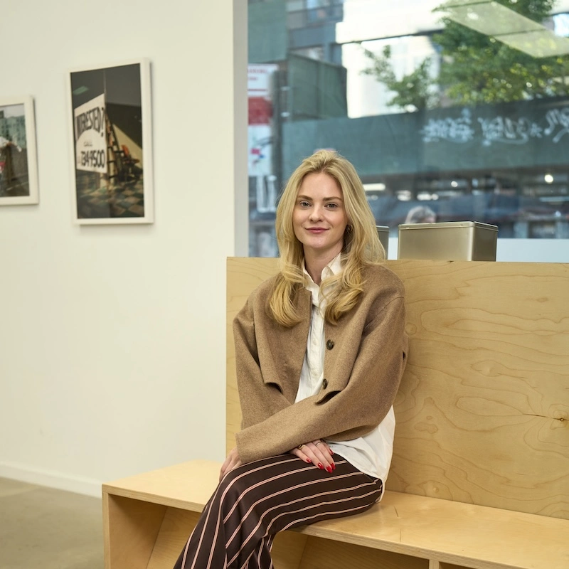 A woman sitting on a wooden bench in an art gallery, wearing a brown cardigan and striped pants, smiling at the camera, with photographs displayed in the background.