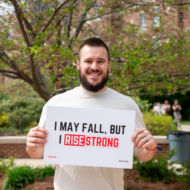Joe Mankowski holding a sign reading "I may fall, but I RiseStrong"