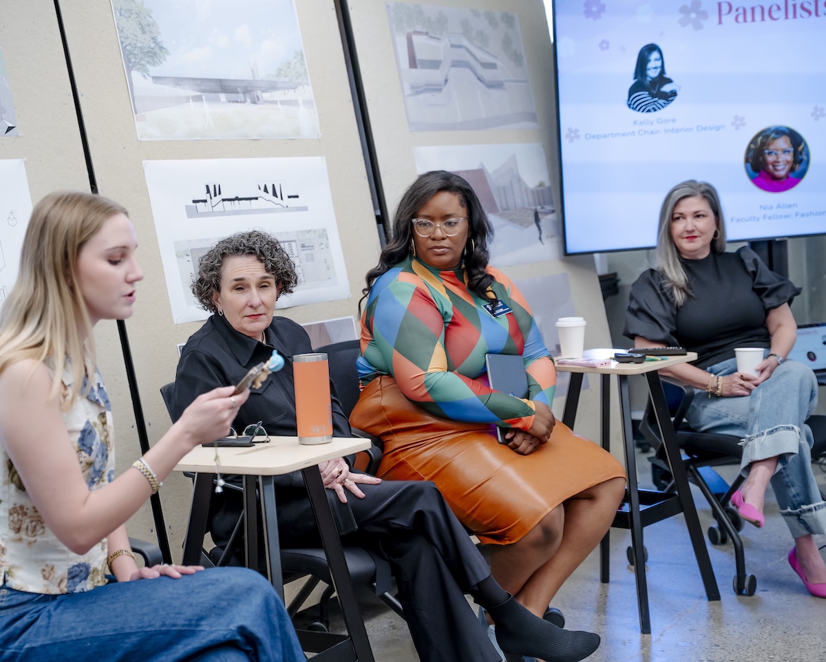 A student moderates a Women’s History Month panel at Belmont’s O’More College, joined by faculty and industry professionals seated in front of design boards and a presentation screen.