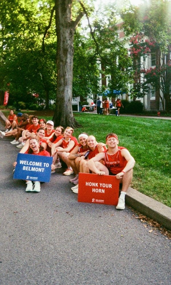TT students pose, sitting on a curb in front of a dorm, welcoming students in. Signs read "Welcome to Belmont" and "Honk your Horn" with the Belmont University logo.