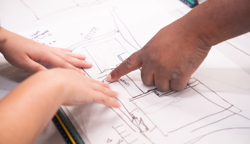 Close-up of three hands over a sheet of paper covered in pencil and marker sketches of a floor plan or structure. One hand points to a detail in the drawing while two smaller hands rest on the paper.