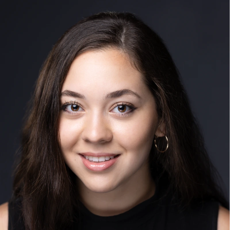 Smiling young woman with long dark hair, wearing a black top and gold hoop earrings, in a professional headshot.
