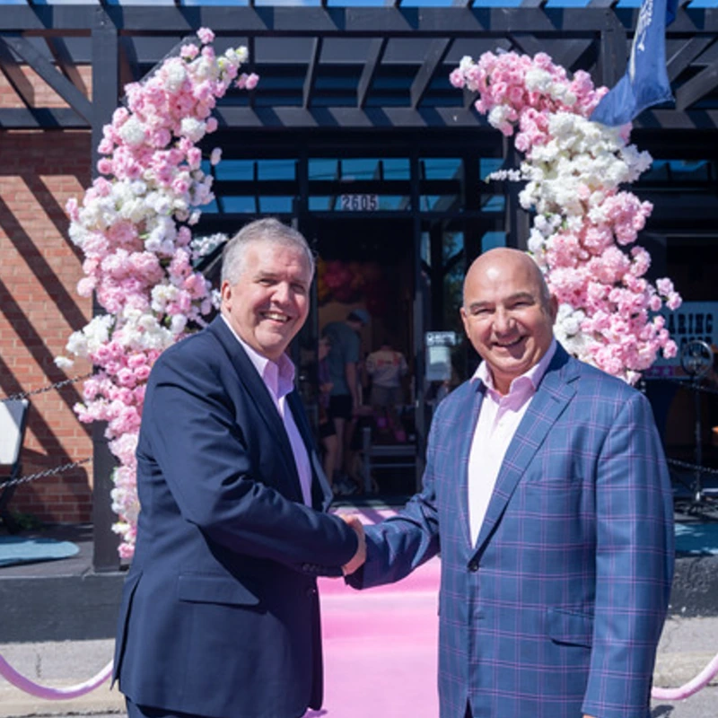 Two men shake hands on a pink carpet with a cherry blossom arch in the background