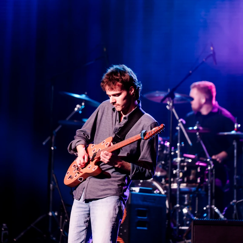Guitarist playing an electric guitar live on stage, illuminated by blue lights, with a drummer behind