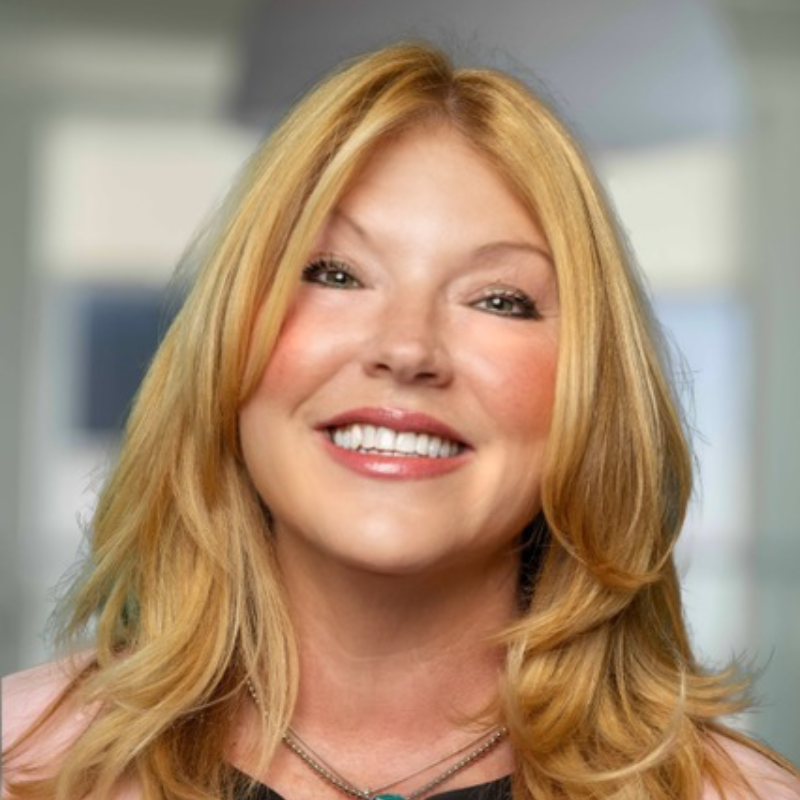 Smiling woman with long blonde hair wearing a pink blazer, against a background of framed portraits.