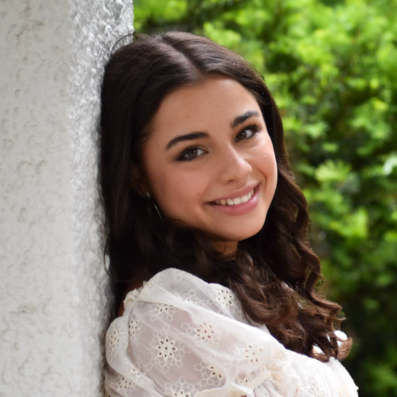 Portrait of a happy young woman with dark wavy hair wearing a white top, smiling outdoors against a textured wall.