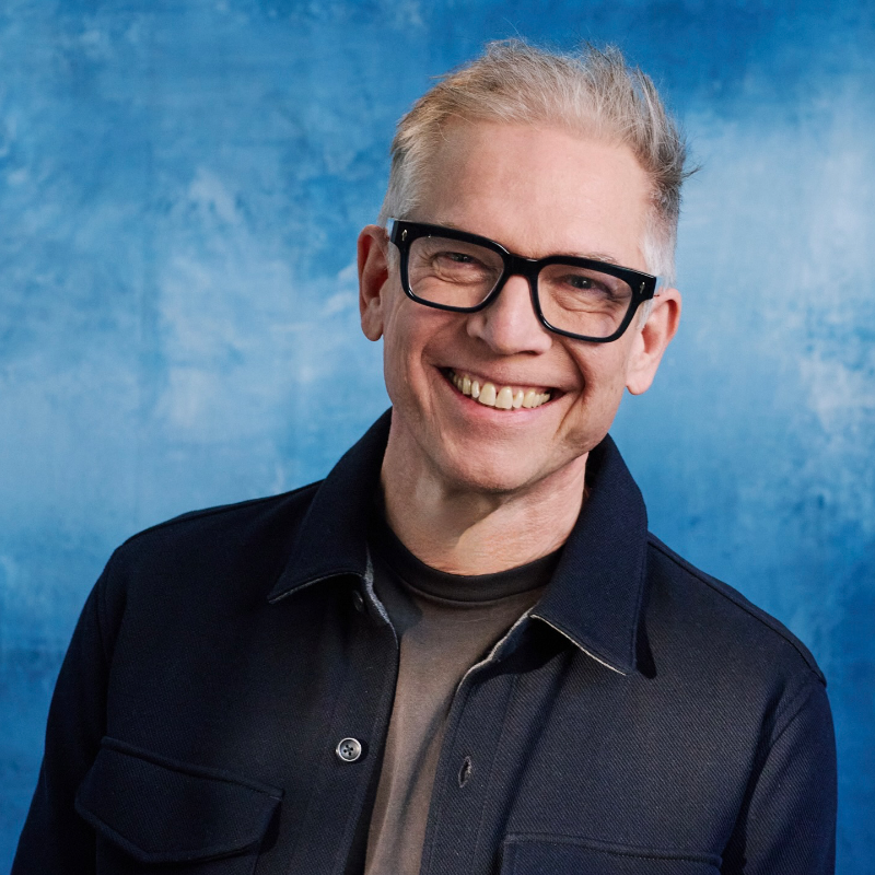 A smiling man with short grey hair and black glasses wears a dark jacket against a blue background.