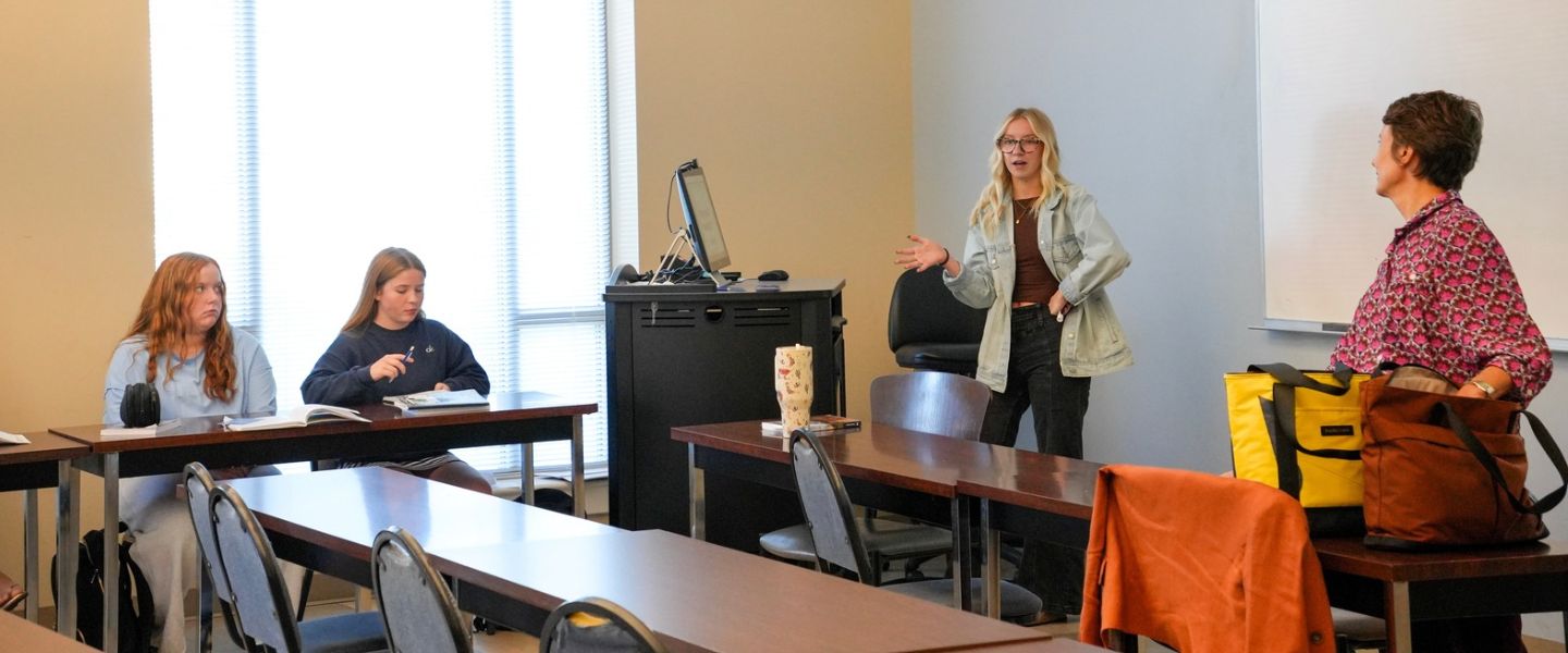 Mentor Jordyn Santos instructs class. Girl standing up in front of white board giving presentation. Students listening. 