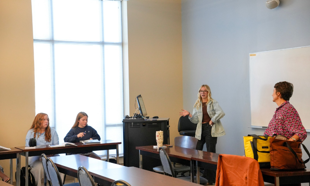 Jordyn Santos instructs course. She is standing up in front of the class gesturing to the board and three students are watching her.