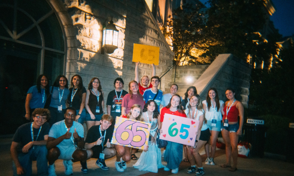 TT group poses at night together in front of building. three people hold colorful signs that say "65"