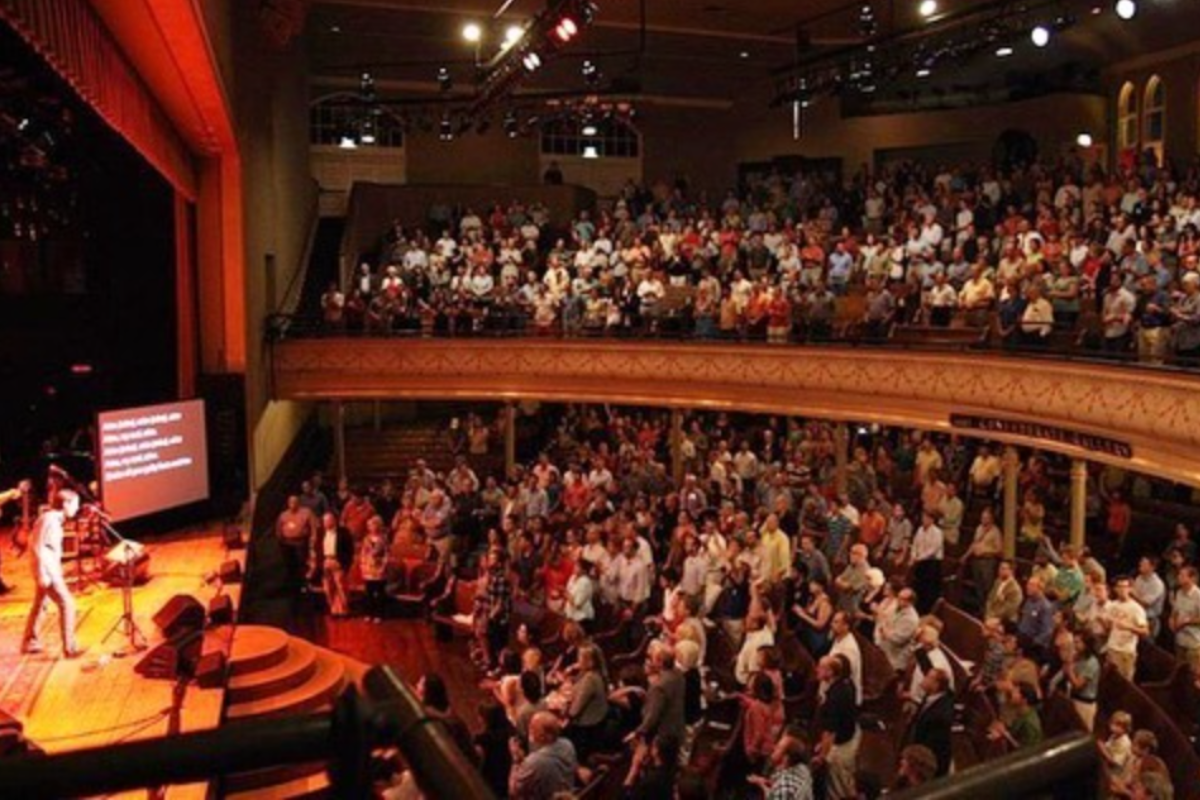 Auditorium packed with an audience watching a performer on stage with a large projection screen.