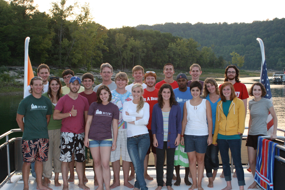 Smiling group of young adults, some wearing 'RUP' shirts, standing on a boat with a lake and forested hills.