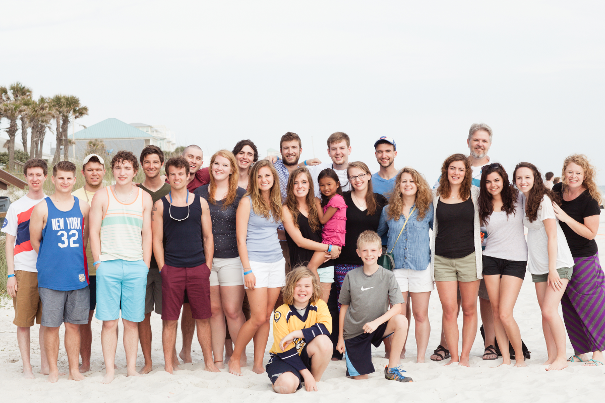 Large group of smiling people, including adults and children, posing on a sandy beach with palm trees in the background.