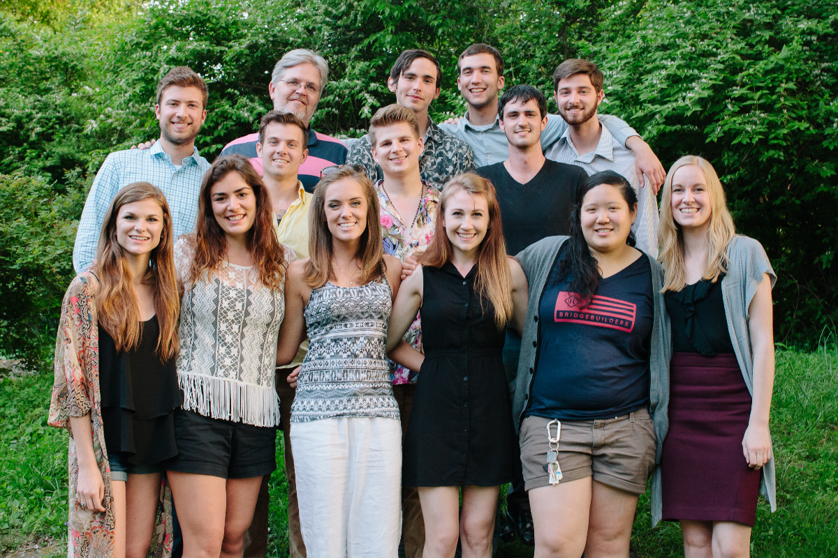 Group photo of diverse young adults and older individuals smiling outdoors with green foliage in the background.