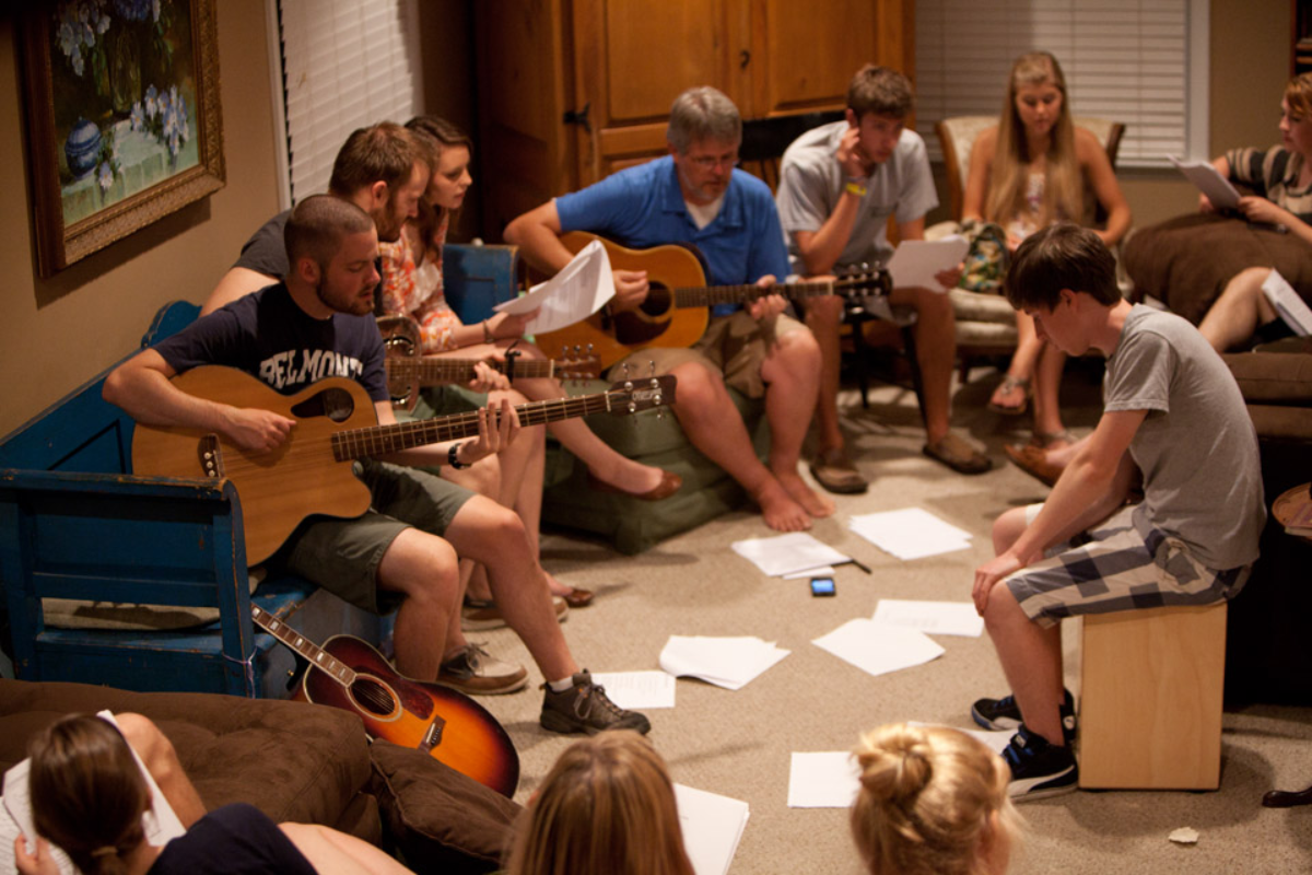 A group of young adults plays acoustic guitars and a cajon during a casual music session indoors.