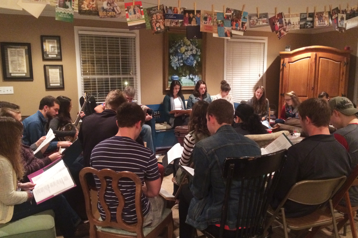 Young adults attend a community group meeting, reading handouts in a cozy, informal living room setting.