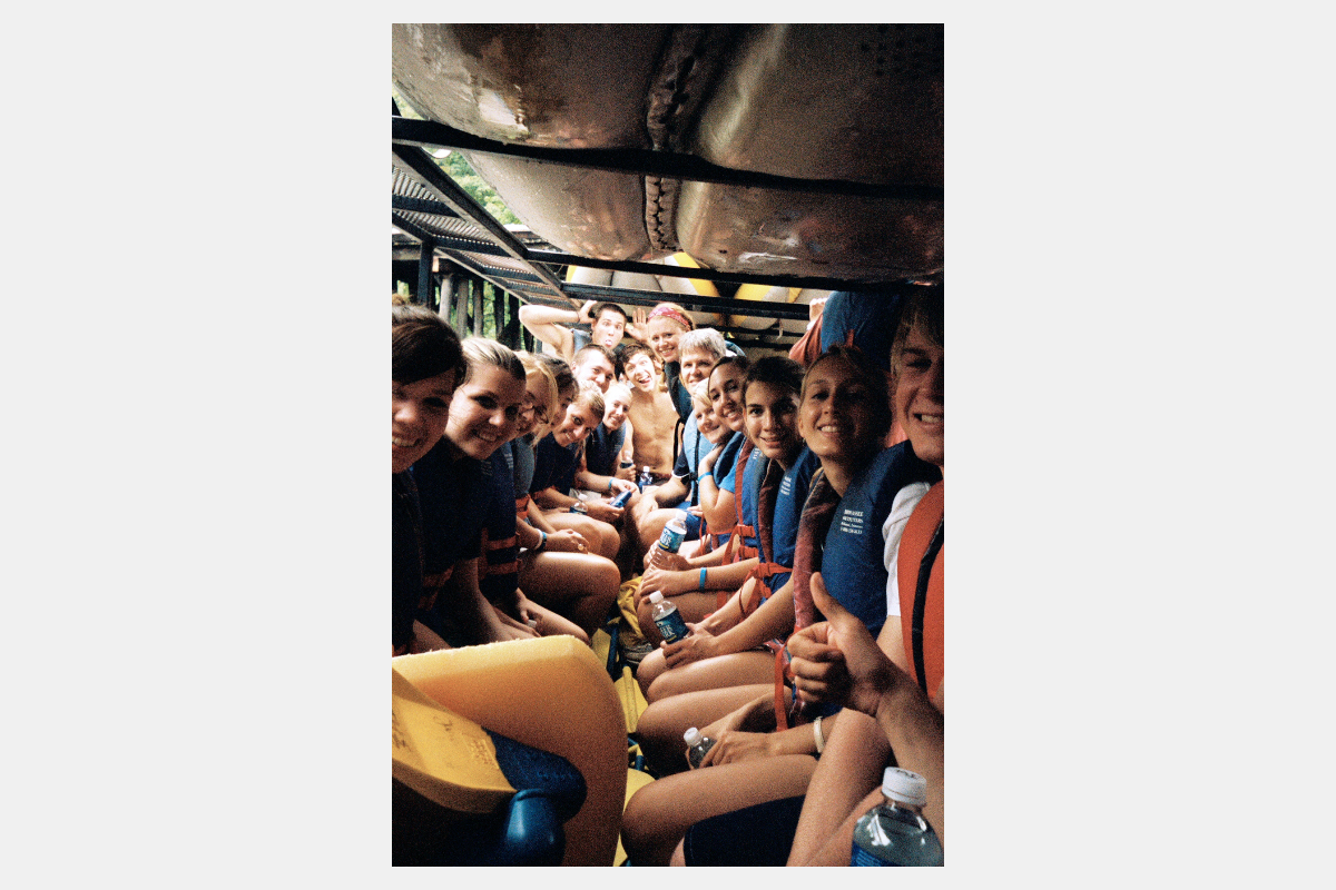 Group of smiling teenagers in life vests packed in a transport vehicle with canoes overhead, ready for a water adventure.