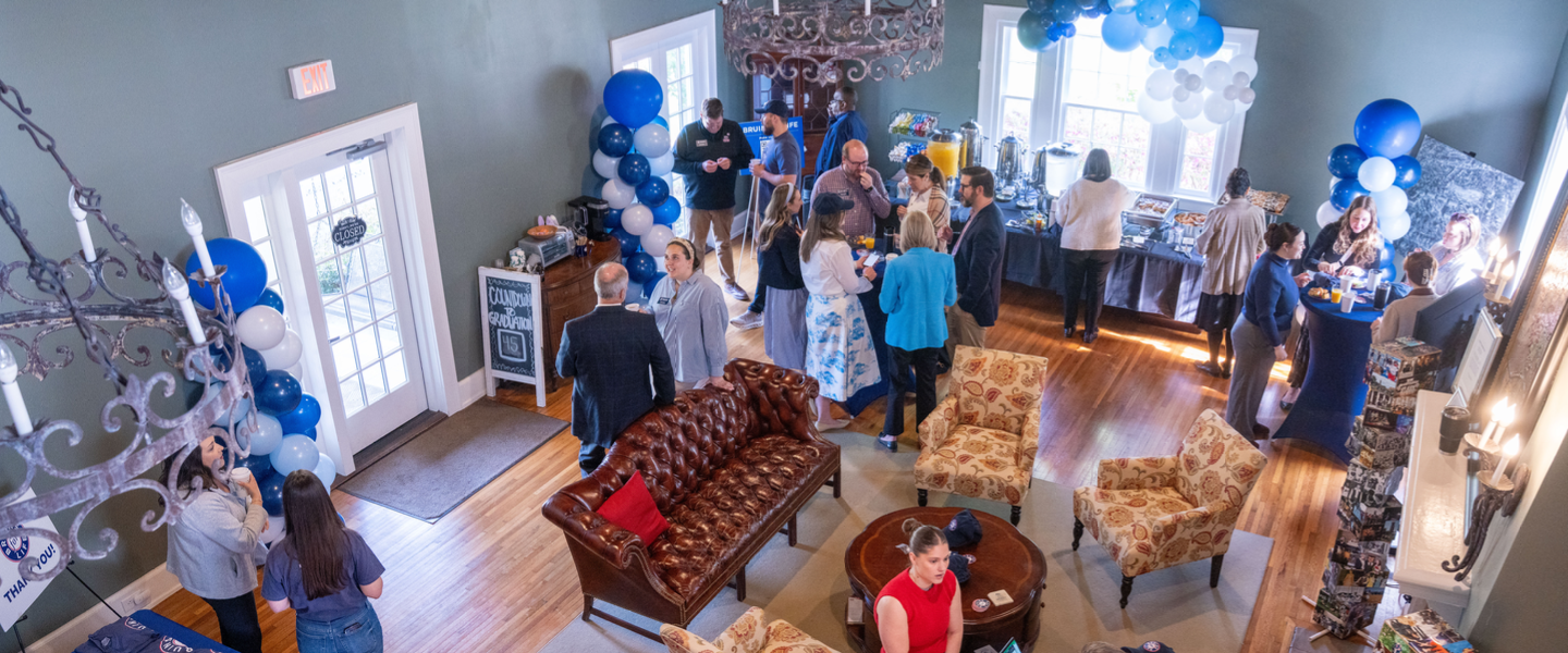 Guests enjoying a celebratory indoor social event with food stations, blue and white balloons, and casual seating.