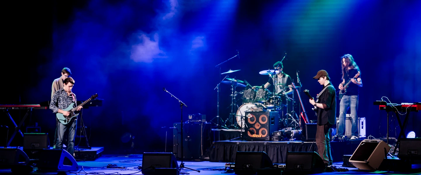 A live band performs on stage under vibrant blue lighting, with guitarists, a drummer and keyboards.