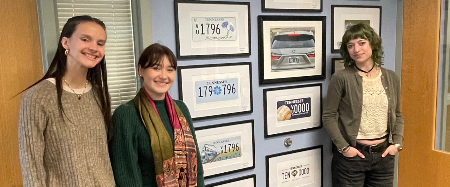 Three smiling women, one holding a Certificate of Appreciation, at an event with license plate designs on display.